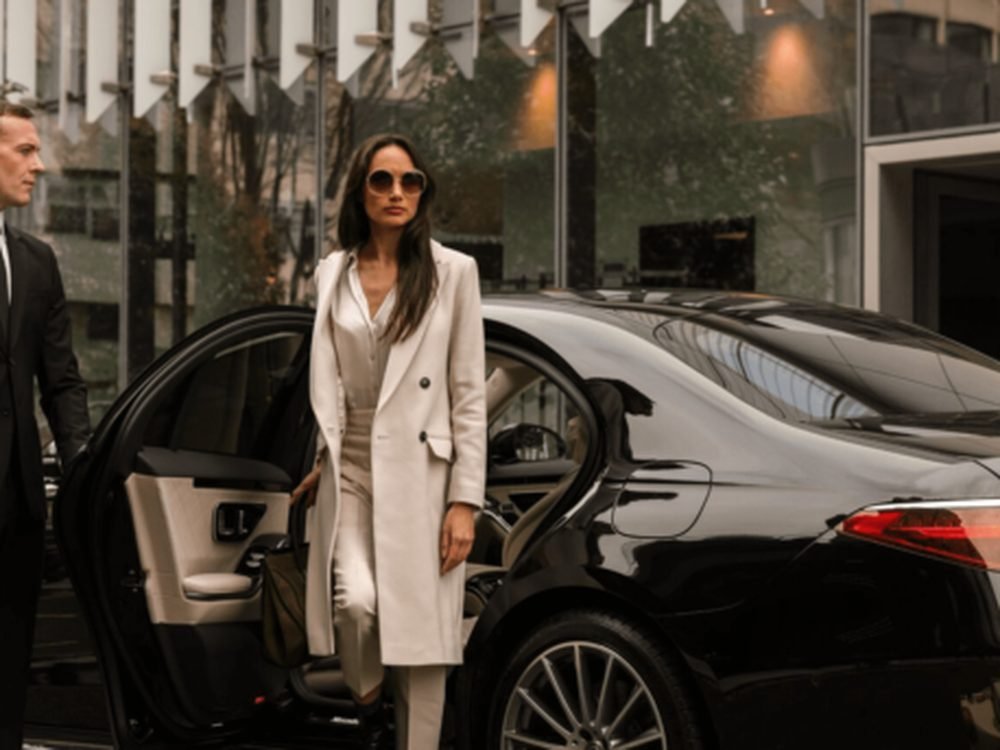 A chauffeur holds the door as a client arrives at her destination in a black chauffeured sedan.