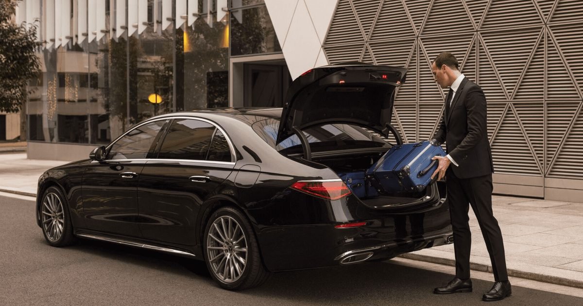 A chauffeur loading a guest's luggage into the trunk of a black chauffeured sedan.